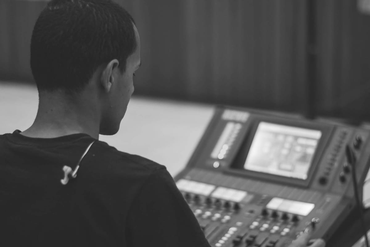 Black-and-white over-the-shoulder of Victor at a digital mixing console with an onboard touchscreen, earphones resting around his neck.