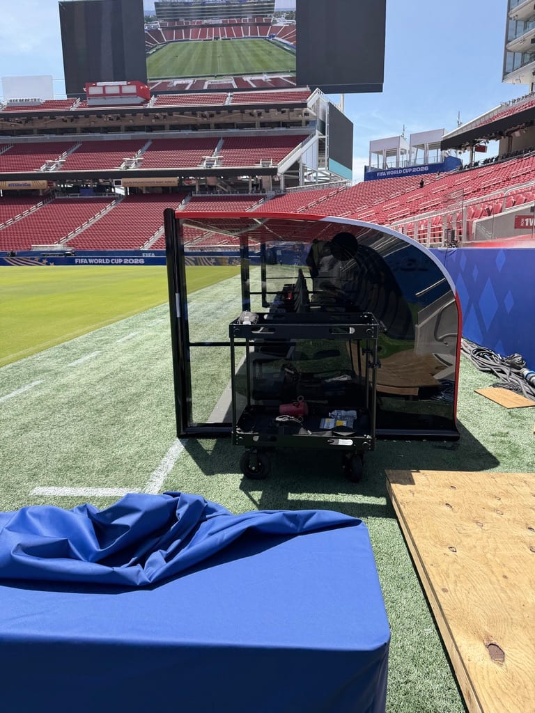 FIFA World Cup 2026 — a covered pitchside broadcast/commentary position in the stadium bowl, empty red seats and the green pitch beyond, tournament boards around the stands.