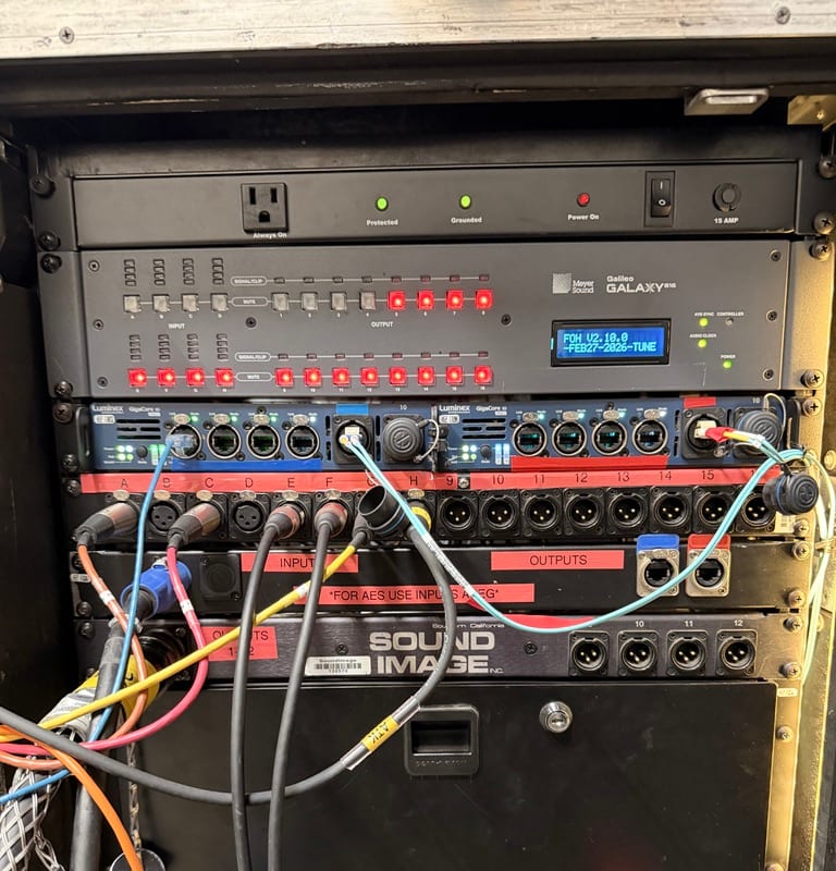 Meyer Sound Galileo amplifier rack — rows of amp faces and patch panels in a road case, status LEDs glowing in low backstage light.