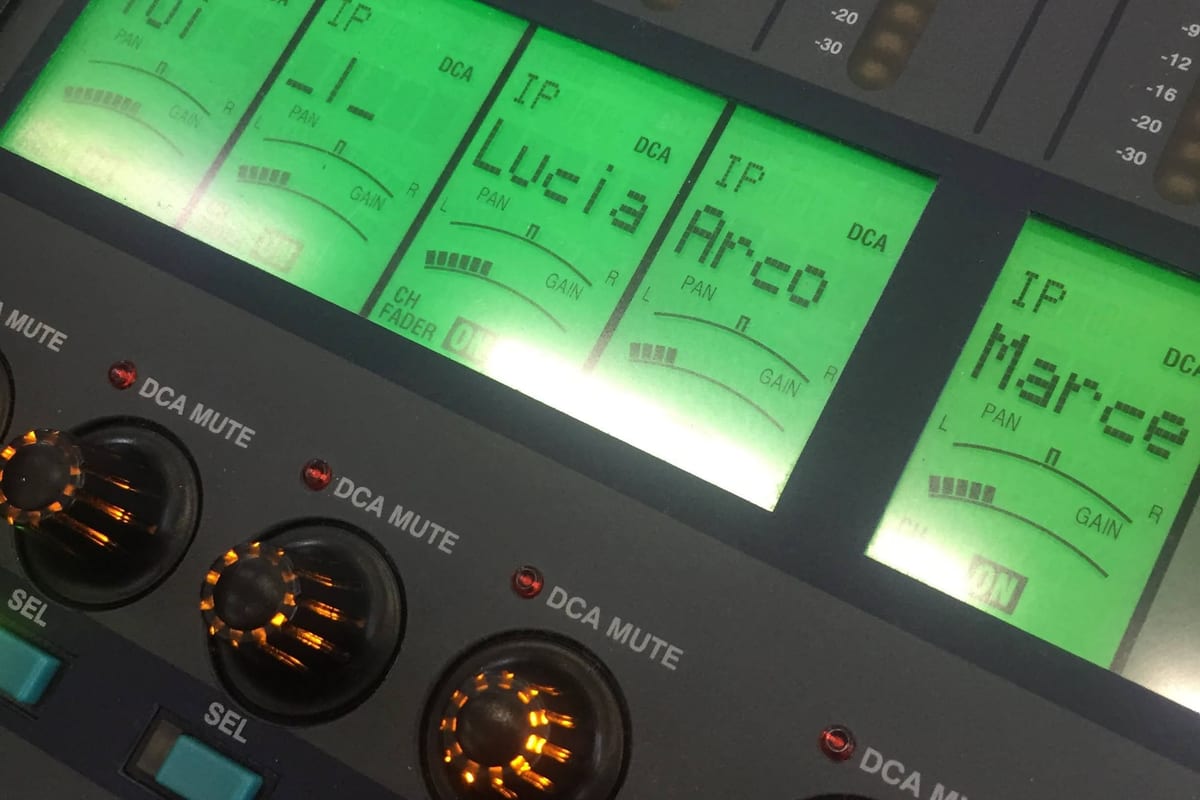 Macro of a Yamaha PM5D channel strip — bright lime-green LCD scribble strips spell 'Tui / Lucia / Arco / Marce' beside meter ladders, orange-glow rotary encoder rings below.