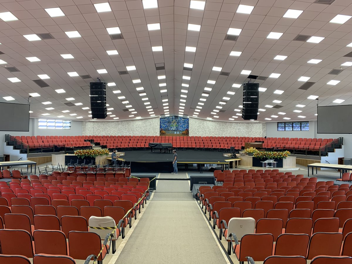 The UNASP auditorium before doors — banked red seating, flown line arrays over the room, a grand piano on the stage under a dense ceiling lighting grid.